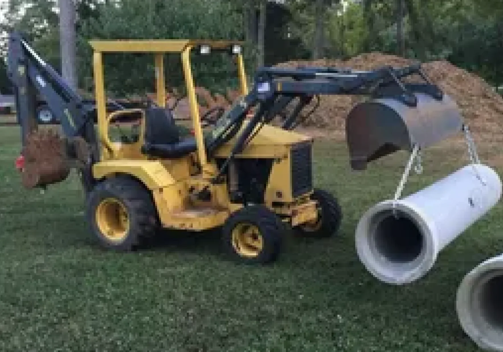 Small yellow backhoe moving a large pipe outdoors.